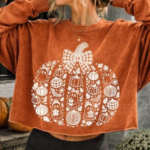 Könnte beinhalten: Ein langärmeliges Sweatshirt in Rostorange mit einem großen weißen Grafikdruck in Kürbisform, der aus kleineren Kürbissen, Geistern und Blättern besteht. Das Kürbismotiv ist mit einer großen Vichy-Schleife verziert und hat einen offenen Saum.