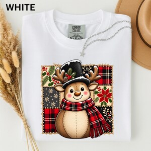 Könnte beinhalten: Ein weißes T-Shirt mit einer Grafik eines Cartoon-Rentiers, das einen schwarzen Zylinder und einen rot-schwarz karierten Schal trägt. Das Rentier ist in einem quadratischen Patchwork-Design mit Mustern aus rotem Schottenkaro, Schneeflocken und Weihnachtssternen zentriert.