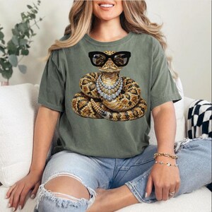 Puede incluir: Una camiseta de manga corta color verde salvia con un estampado gráfico de una serpiente enroscada marrón y beige que lleva gafas de montura negra y un collar de varias capas de pedrería plateada, centrado en el pecho de la prenda.
