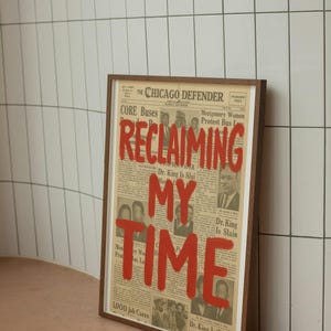 Puede incluir: Impresión enmarcada con un fondo de periódico vintage. El titular dice "The Chicago Defender". Texto rojo grande que dice "RECLAIMING MY TIME". La impresión está en un marco de madera marrón, contra una pared de azulejos blancos.