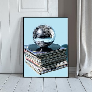 Puede incluir: Una impresión enmarcada que presenta una bola de discoteca plateada encima de una pila de discos de vinilo. El fondo es azul claro y el marco es negro. Los discos tienen fundas coloridas, y la imagen está sobre una puerta y un suelo blancos.