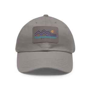 Puede incluir: Gorra de béisbol gris con visera curva. La gorra presenta un parche rectangular con un diseño de montaña y sol en azul, naranja y turquesa. El texto "NATURE'S ADVENTURES" está impreso en el parche.