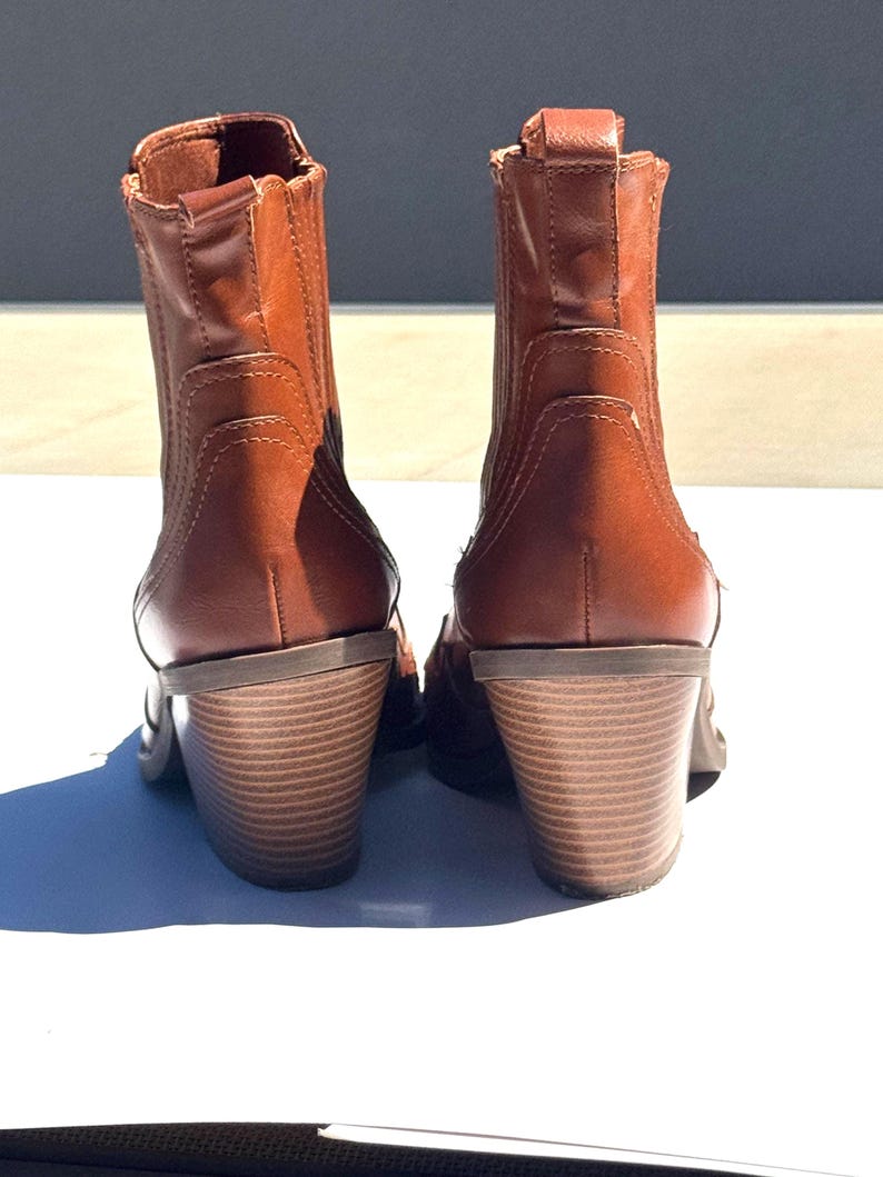May include: A pair of brown leather ankle boots with stacked heels. The boots have a classic design with a rounded toe and elastic side panels. The heels are approximately 3 inches high, providing a stylish lift.