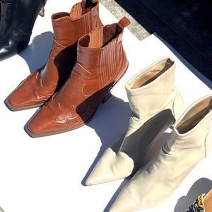 May include: An assortment of ankle boots in various colors. Two brown leather boots with decorative stitching and a square toe are next to a pair of cream-colored pointed-toe boots. A black boot is also visible.