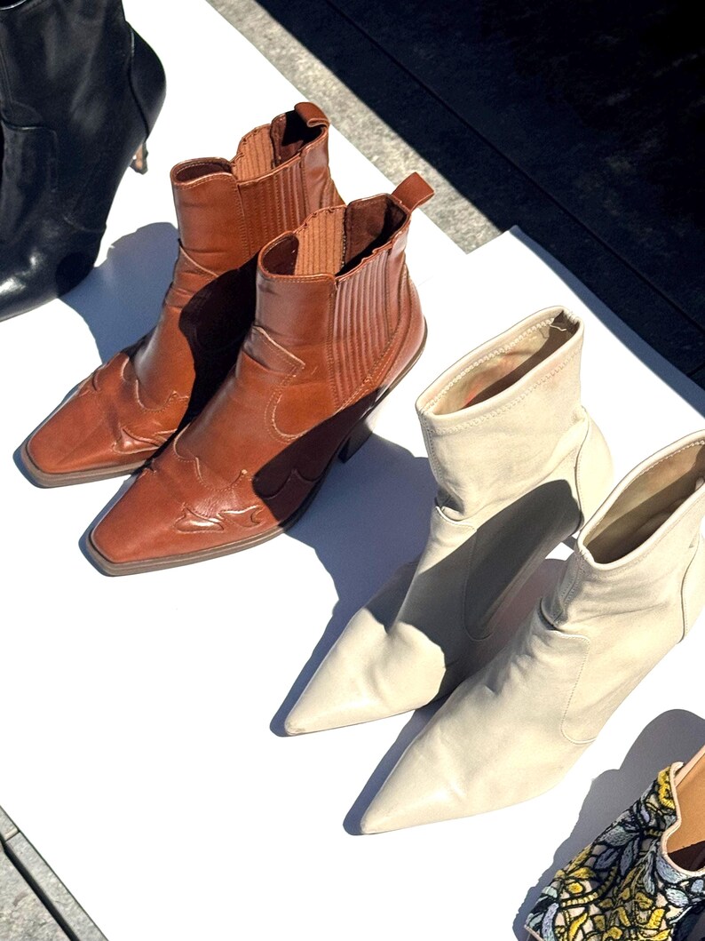 May include: An assortment of ankle boots in various colors. Two brown leather boots with decorative stitching and a square toe are next to a pair of cream-colored pointed-toe boots. A black boot is also visible.