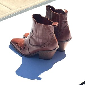 May include: A pair of brown leather ankle boots with a Western-inspired design. The boots have a pointed toe, decorative stitching, and a stacked heel. The boots are approximately 6 inches tall.