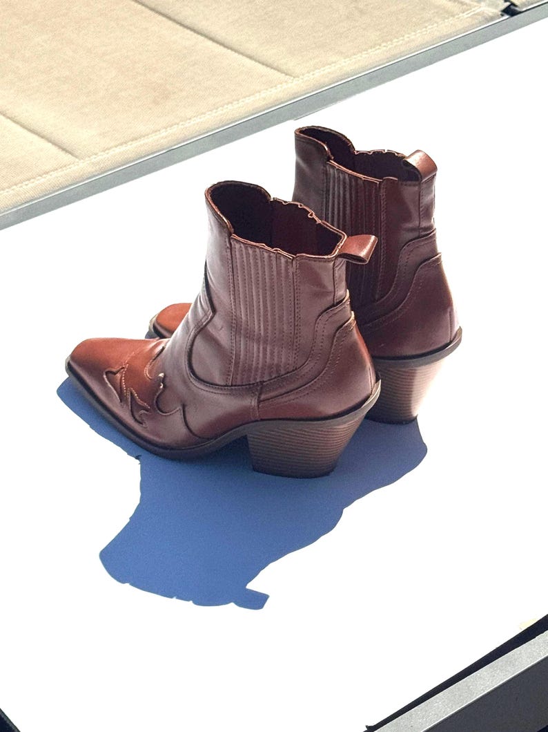May include: A pair of brown leather ankle boots with a Western-inspired design. The boots have a pointed toe, decorative stitching, and a stacked heel. The boots are approximately 6 inches tall.