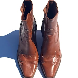 May include: A pair of brown leather ankle boots with a Western-inspired design. The boots have a pointed toe, decorative stitching, and a pull tab at the top. The boots are approximately 8 inches tall.