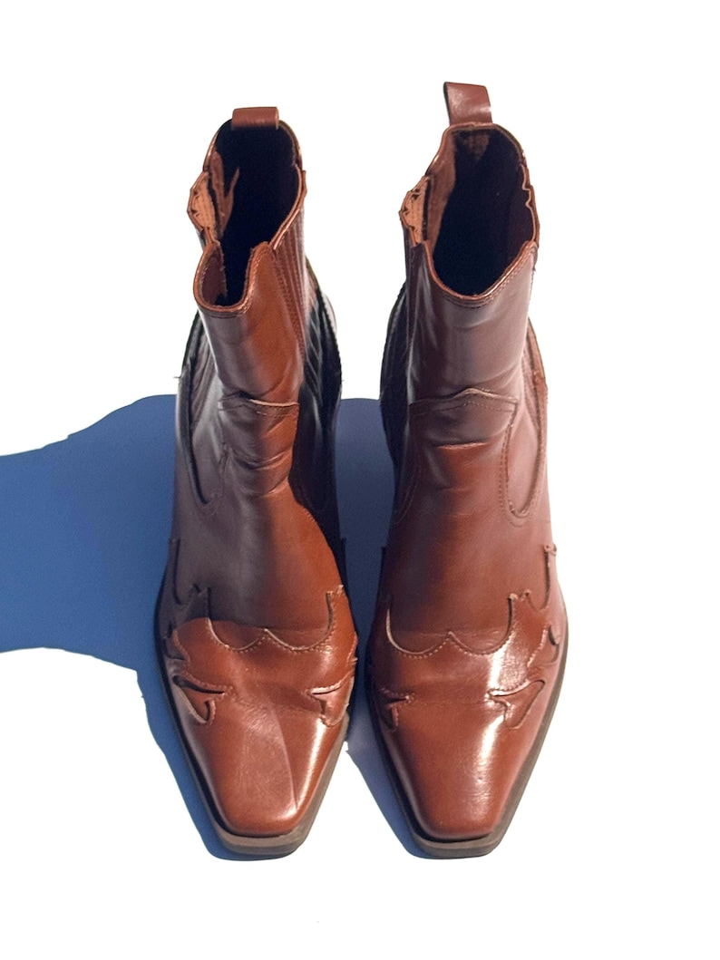 May include: A pair of brown leather ankle boots with a Western-inspired design. The boots have a pointed toe, decorative stitching, and a pull tab at the top. The boots are approximately 8 inches tall.