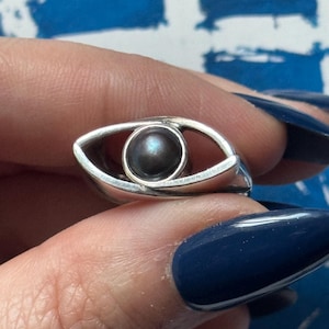 May include: A silver ring featuring an eye design. The ring has a polished silver band with an eye-shaped setting. A dark, iridescent pearl is set in the center of the eye. The ring is held up against a blue background.