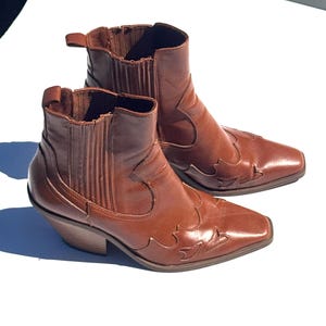 May include: A pair of brown leather ankle boots with a Western-inspired design. The boots feature a pointed toe, decorative stitching, and a stacked heel. The pull tabs and elastic side panels add to the boot's style.