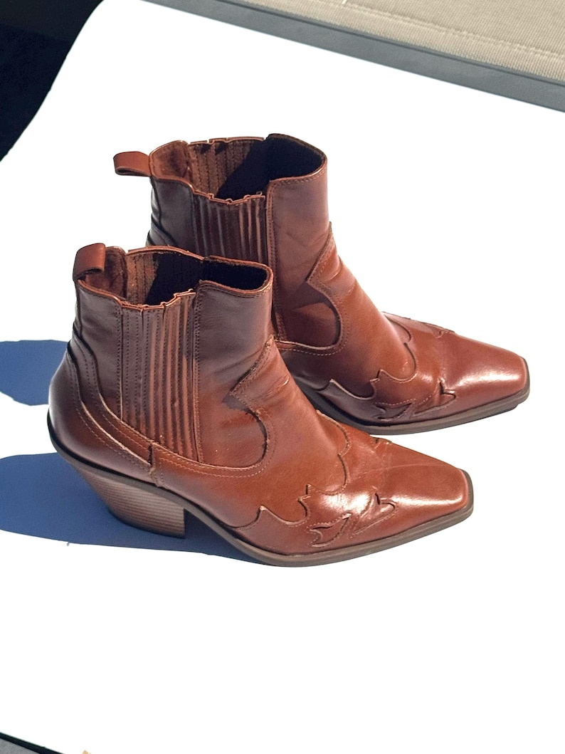 May include: A pair of brown leather ankle boots with a Western-inspired design. The boots feature a pointed toe, decorative stitching, and a stacked heel. The pull tabs and elastic side panels add to the boot's style.