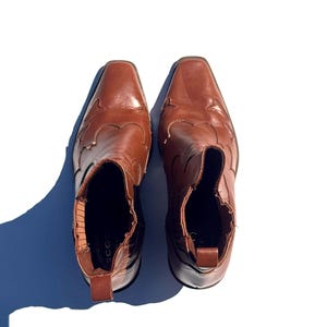 May include: A pair of brown leather ankle boots with a square toe and decorative stitching on the vamp. The boots have a pull tab at the back and a low heel. The boots are approximately 8 inches tall.