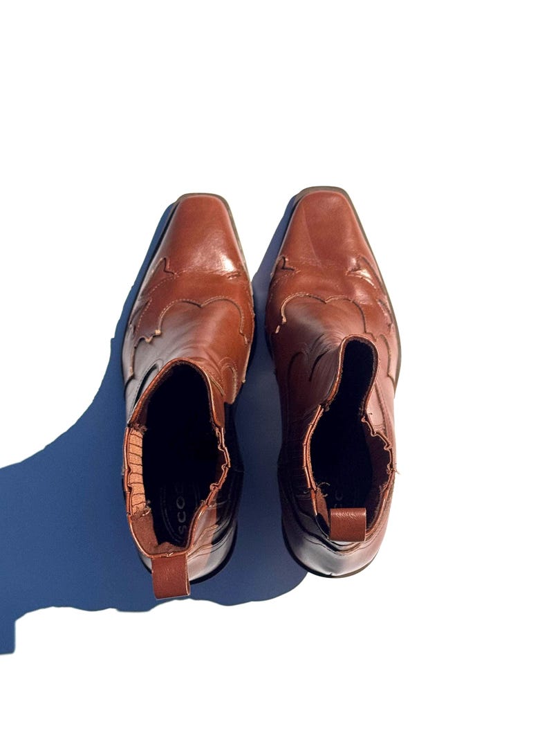 May include: A pair of brown leather ankle boots with a square toe and decorative stitching on the vamp. The boots have a pull tab at the back and a low heel. The boots are approximately 8 inches tall.