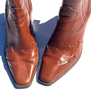 May include: A pair of brown leather cowboy boots. The boots have a square toe and decorative cutouts on the vamp. The shaft is mid-height, and the leather has a polished finish.