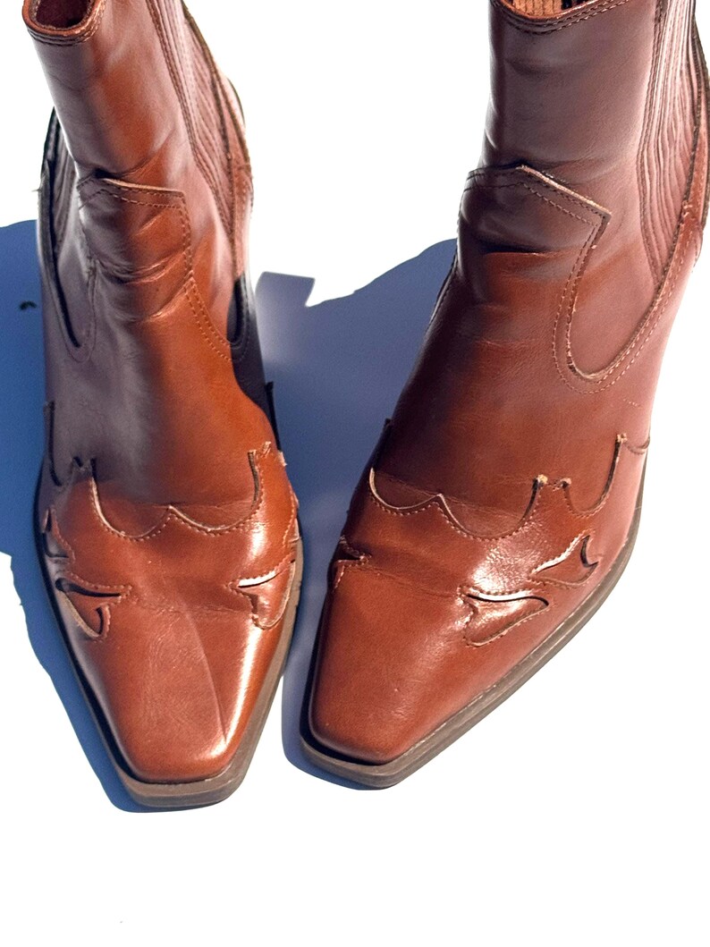 May include: A pair of brown leather cowboy boots. The boots have a square toe and decorative cutouts on the vamp. The shaft is mid-height, and the leather has a polished finish.