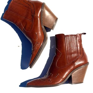 May include: A pair of brown leather ankle boots with a pointed toe and a stacked wooden heel. The boots have decorative stitching and a pull tab at the top. The heels are approximately 3 inches high.