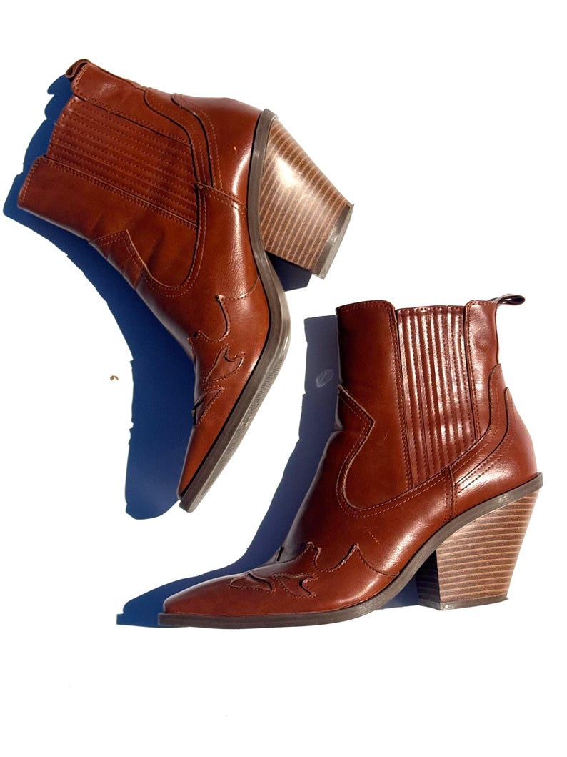 May include: A pair of brown leather ankle boots with a pointed toe and a stacked wooden heel. The boots have decorative stitching and a pull tab at the top. The heels are approximately 3 inches high.
