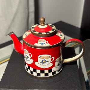 May include: A red decorative teapot with a lid, spout, and handle. The teapot is adorned with images of coffee cups and saucers. The base features a black and white checkered pattern. The lid is topped with a small, round, gold-colored knob.