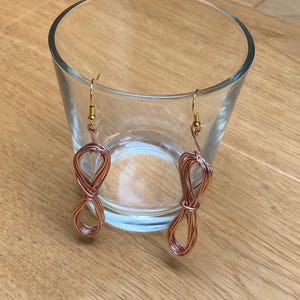 Peut inclure: Une paire de boucles d'oreilles en fil de cuivre de couleur cuivrée, en forme de symbole infini. Les boucles d'oreilles sont suspendues à des crochets dorés. Elles sont présentées devant un verre transparent sur une surface en bois.