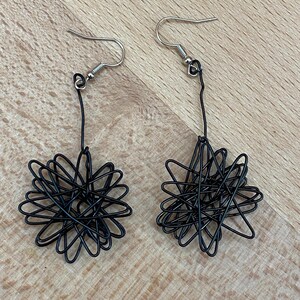 Peut inclure: Une paire de boucles d'oreilles en fil noir. Chaque boucle d'oreille présente un motif en forme d'éclat d'étoile fait de fil bouclé, suspendu à un crochet d'oreille argenté. Les boucles d'oreilles sont présentées sur une surface en bois brun clair.