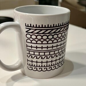 Könnte beinhalten: Weiße Keramik-Tasse mit einem schwarzen geometrischen Muster. Das Design zeigt verschiedene Formen, darunter Linien, Kreise und Kurven. Die Tasse hat einen gebogenen Griff.