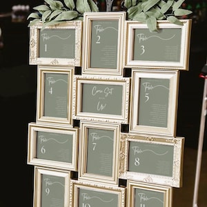 Puede incluir: Un plano de asientos de boda con once tarjetas enmarcadas que muestran los números de mesa y los nombres de los invitados. Los marcos ornamentados de color crema están dispuestos en una cuadrícula. Cada tarjeta tiene un fondo verde apagado con una elegante escritura blanca. El texto "Come Sit With Us!" está en el centro.