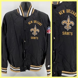 Peut inclure: Veste noire des Saints de la Nouvelle-Orléans avec des détails dorés et blancs. Elle présente le logo de l'équipe et l'inscription "New Orleans Saints" en or. Comprend un écusson NFL. Fermeture boutonnée et bordure rayée.