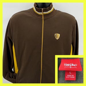 May include: Brown zip-up jacket with yellow accents on the sides and a striped collar. Features a gold emblem on the chest and a red tag with the brand name and size.