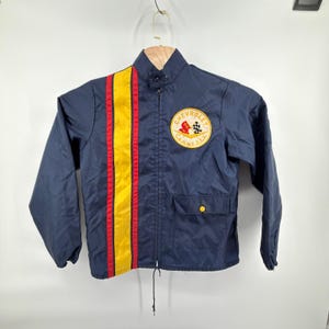 May include: A navy blue zip-up jacket featuring a vertical yellow stripe flanked by two thin red stripes on the left side. A circular Chevrolet Corvette logo patch is on the upper right chest. The jacket includes a single front pocket with a yellow snap.