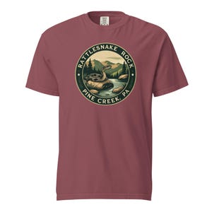 以下が含まれることがあります： マルーン色のTシャツ。円形のグラフィックには「Rattlesnake Rock Pine Creek, PA」の文字と、川の中の岩の上にいるヘビと山の背景が描かれています。柔らかい素材で作られたTシャツです。