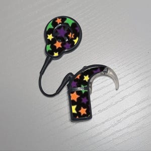 May include: A cochlear implant processor and its external coil connected by a thin black cable. Both components feature a black surface decorated with a repeating pattern of small, multi-colored stars in shades of orange, yellow, green, and purple.