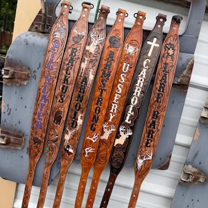 Puede incluir: Siete correas de cuero para rifle grabadas a medida dispuestas verticalmente. Cada correa presenta intrincados tallados florales, imágenes de vida silvestre y nombres personalizados como PIG SLAYER, BACKUS, ARNOLD, ARMSTRONG, USSERY, CARROLL y LARE BARE.