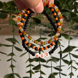Può includere: Braccialetti di perline nere, arancioni, gialle e trasparenti. Un braccialetto recita "BOO" con perline di lettere bianche. I braccialetti sono tenuti in mano su uno sfondo di foglie verdi.