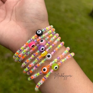 May include: A collection of colorful beaded bracelets, each featuring a central evil eye charm. The bracelets are adorned with a variety of small, vibrant beads in shades of pink, orange, green, purple, and yellow. The bracelets are stacked on a wrist.