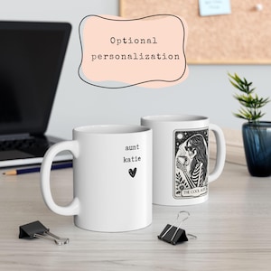 May include: Two white ceramic mugs. One mug has the text "aunt katie" and a heart. The other mug has a black and white illustration of a skeleton with sunglasses holding a cup, with the text "THE COOL AUNT". The background includes a laptop and a plant.