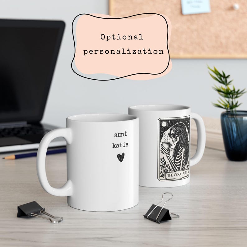 May include: Two white ceramic mugs. One mug has the text "aunt katie" and a heart. The other mug has a black and white illustration of a skeleton with sunglasses holding a cup, with the text "THE COOL AUNT". The background includes a laptop and a plant.