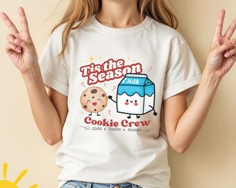 May include: A cream-coloured t-shirt with a festive design. The text reads "Tis the Season" and "Cookie Crew." The graphic features a smiling chocolate chip cookie and a milk carton. The shirt is worn by a person.