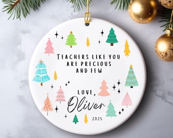 May include: A white ceramic Christmas ornament with a gold hanging loop. The ornament features colorful Christmas trees and the text "Teachers like you are precious and few. Love, Oliver 2025."