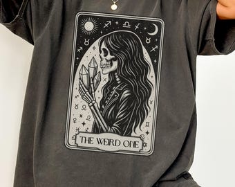 May include: Dark grey t-shirt featuring a tarot card design. The graphic depicts a skeleton with long hair holding crystals, surrounded by celestial symbols. The text "THE WEIRD ONE" is at the bottom of the card.