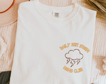 Può includere: T-shirt color crema con la scritta "DAILY SHIT STORM" a semicerchio sopra una nuvola marrone e un fulmine. Le parole "MOMS CLUB" sono sotto.