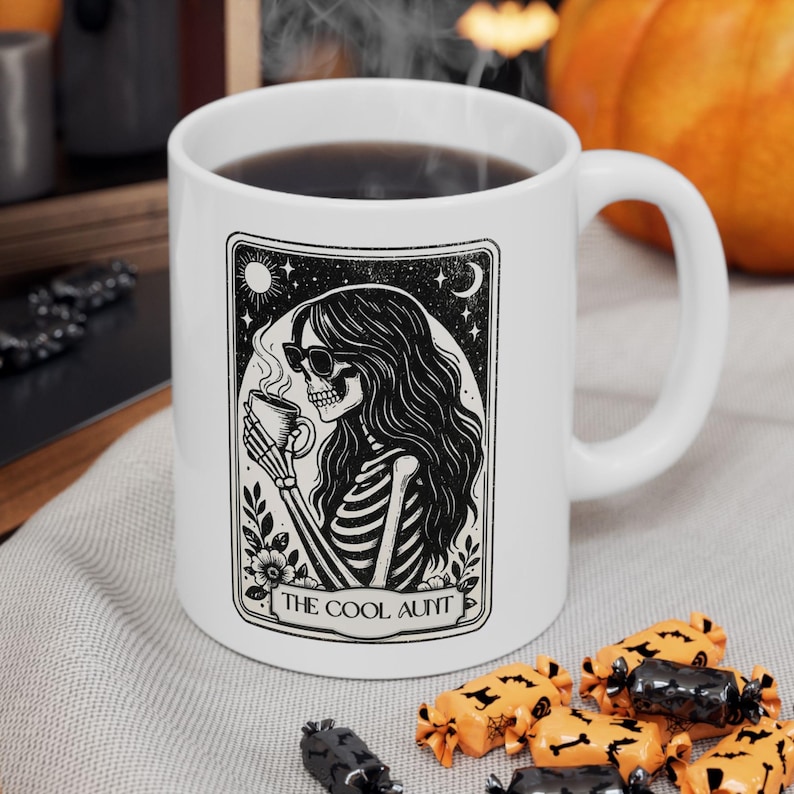 May include: White ceramic mug with a black and white illustration of a skeleton wearing sunglasses and holding a cup of coffee. The text "THE COOL AUNT" is below the illustration. The mug has a curved handle.