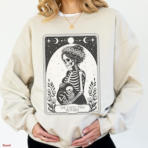 Puede incluir: Sudadera color arena con un gráfico en blanco y negro de una madre esqueleto y un bebé. El diseño incluye elementos florales, lunas y estrellas, con el texto "THE EXPECTING MOTHER".