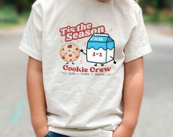 May include: A cream-coloured t-shirt with a graphic of a smiling chocolate chip cookie and a milk carton. The text "Tis the Season Cookie Crew" is printed above the characters. The shirt is worn by a child.