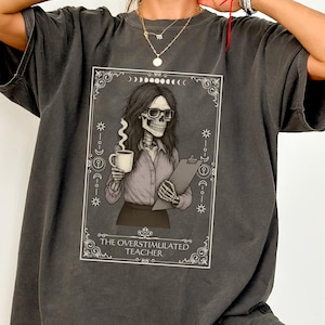 May include: Dark gray t-shirt featuring a framed graphic of a skeleton teacher wearing glasses, holding a coffee mug and clipboard. The graphic includes the text "THE OVERSTIMULATED TEACHER" in a decorative frame. The shirt is a relaxed fit.
