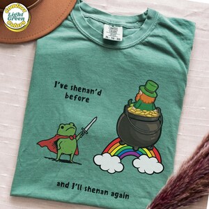 Puede incluir: Camiseta verde claro con el texto "I've shenan'd before and I'll shenan again". La camiseta presenta una rana de dibujos animados con una espada y un duende con una olla de oro sobre un arcoíris. La camiseta está etiquetada como XL.