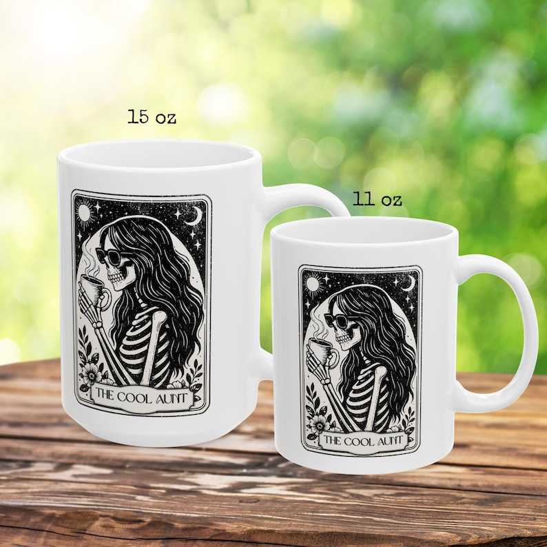 May include: Two white ceramic mugs, 15 oz and 11 oz, with a black and white illustration of a skeleton woman wearing sunglasses and holding a cup. The text "THE COOL AUNT" is below the image. The mugs are on a wooden surface.