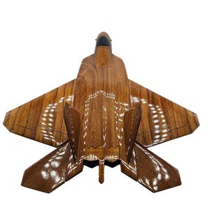 May include: A wooden model of a fighter jet, featuring a detailed design with a brown finish. The wings and body are intricately carved, showcasing the craftsmanship. The jet has a sleek, modern appearance.