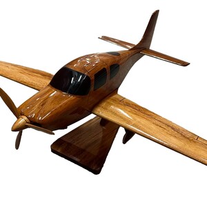 May include: A wooden model airplane with a brown and natural wood finish. The model is on a wooden stand and has a propeller.