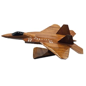 May include: A wooden model of a fighter jet, showcasing a detailed design with a brown finish. The jet features a sleek, aerodynamic shape, including wings, tail fins, and a cockpit. The model is mounted on a wooden stand.